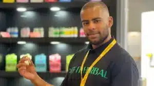 Mann mit DIFF HERBAL T-Shirt und gelbem Namensschildhalter hält kleines gerolltes Produkt vor Regalen.